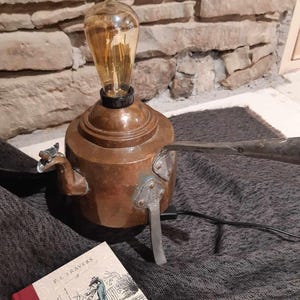 Puede incluir: Una tetera de cobre con una bombilla en la parte superior, utilizada como lámpara de mesa. La tetera tiene un asa y un pico. La lámpara está sentada sobre una alfombra gris oscuro con un libro titulado "Mary Poppins" de P.L. Travers.