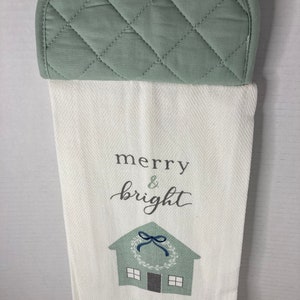 May include: A white kitchen towel with a green and blue house design and the text "merry & bright". The towel has a green quilted pot holder attached to the top.