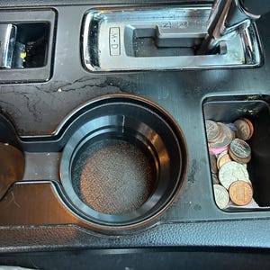 May include: A close-up of a car's center console with a cup holder and a gear shift lever. The gear shift lever has the letters "N", "R", and "W-D" on it. There are coins in the cup holder.