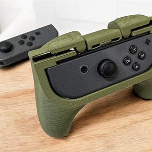 May include: An olive green gaming controller grip with a black center panel and buttons. A separate black controller is visible in the background. The grip has a textured surface and ergonomic design, enhancing the gaming experience.