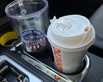May include: A close-up of a car's cup holder with two drinks. The cup holder is made of black plastic and has a silver trim. The drink on the left is in a clear plastic cup with a red, white, and blue "Bless the USA" sticker. The drink on the right is in a white paper cup with a red and orange "Dunkin'" logo.