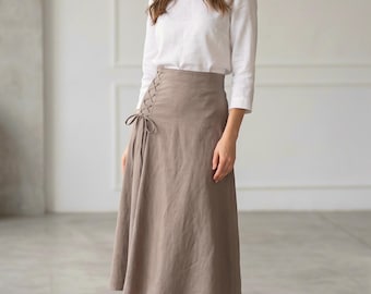 May include: A woman wearing a white long-sleeved shirt and a taupe-colored midi skirt. The skirt features a lace-up detail on the side. The outfit is completed with nude-colored heels. The setting is a well-lit room with a neutral background.