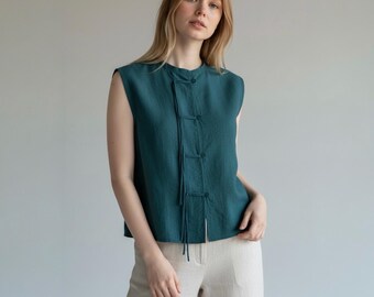 May include: Teal sleeveless top with a mandarin collar and decorative string ties. The top has three fabric-covered buttons and is paired with light beige pants. The outfit is made of a lightweight fabric.