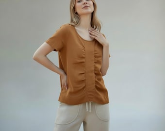 May include: A short-sleeved, rust-colored top with a scoop neckline and vertical gathering detail down the center. Paired with light beige pants with front pockets and a drawstring waist. The outfit is casual and comfortable.