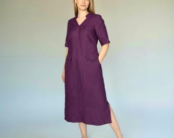 May include: A plum-colored linen dress with short sleeves and a V-neck. The dress features two front pockets and a side slit. The model is wearing the dress. The dress is knee-length.
