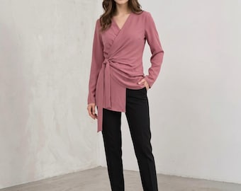 May include: A long-sleeved, dusty rose wrap blouse featuring a V-neckline and a side-tie closure, paired with slim-fit black trousers. The blouse has a structured collar and a draped front panel that extends to the hip, while the trousers are tailored.