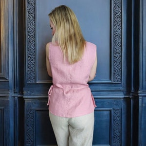 Pode incluir: Uma mulher vestindo uma blusa de linho rosa sem mangas com cintura amarrada e cal&ccedil;as de linho bege. A blusa tem um caimento solto e um comprimento ligeiramente curto. As cal&ccedil;as s&atilde;o de perna reta e t&ecirc;m cintura alta.