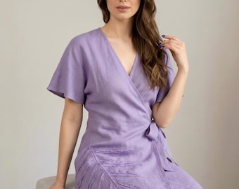 May include: A lavender wrap dress with short sleeves and a V-neck. The dress features a leaf design on the front and a tie closure at the waist. The dress is made of a lightweight fabric.