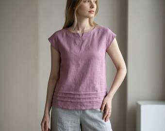 May include: A mauve-colored linen top with a V-neckline and short sleeves. The top features horizontal pleats at the waist and is paired with gray linen culottes. The outfit is simple and elegant, suitable for casual wear.