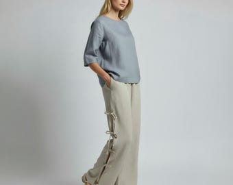 May include: A woman wearing a light blue top with three-quarter sleeves and beige wide-leg pants. The pants have decorative ties on the side. The outfit is made of a lightweight fabric, suitable for warm weather.