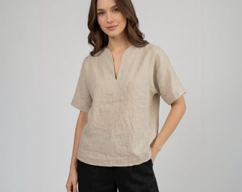 May include: A beige linen top with a V-neckline and short sleeves. The top is paired with black pants. The fabric has a textured appearance. The model is standing in front of a plain, light-colored background.