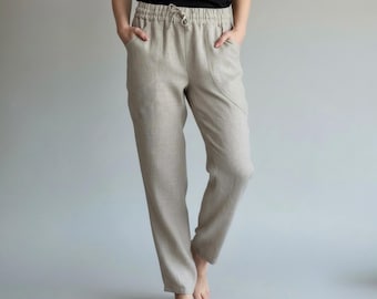 May include: Beige linen pants with a drawstring waist and pockets. The pants have a relaxed fit and a neutral color, suitable for casual wear. The model is wearing a black top.