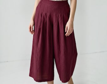 May include: Deep burgundy wide-leg trousers featuring a high-waisted design with vertical pleats at the front. The pants have a loose, cropped silhouette that falls below the knee, paired with a simple, light-colored sleeveless top tucked in.