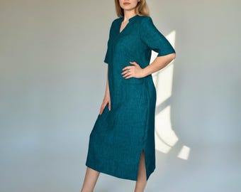 May include: Teal-colored linen dress with a V-neckline and short sleeves. The dress features a single pocket and a side slit. The model is standing in front of a plain background. The dress is knee-length.