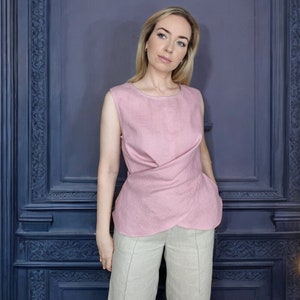 Pode incluir: Uma mulher vestindo uma blusa de linho rosa com um detalhe de n&oacute; e cal&ccedil;as de linho bege. A blusa tem um decote redondo e &eacute; sem mangas.