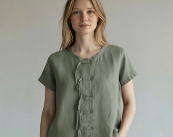 May include: A short-sleeved, olive green linen top with a round neckline and a series of fabric-covered buttons down the front. The top has a relaxed fit and is made of a lightweight, breathable fabric, suitable for warm weather.