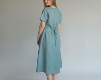 May include: A light teal linen dress with short sleeves and a tie-back waist. The dress has a simple, elegant design and falls to mid-calf length. The model is facing away from the camera, with a neutral background.