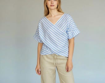 May include: A light blue and white striped wrap top with short sleeves and a V-neck. The top is paired with tan pants. The sleeves have a ruffled detail. The clothing is made of a lightweight fabric.