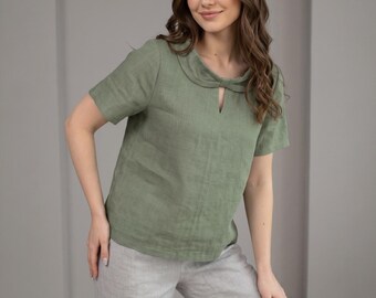 May include: A woman wearing a short-sleeved, olive green linen top with a unique knotted neckline and a small keyhole detail. She is also wearing light gray linen pants. The outfit is casual and stylish.