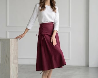 May include: A woman wearing a white long-sleeve top and a burgundy midi skirt with a lace-up detail. The skirt is a solid color and has a flowing silhouette. The woman is wearing nude heels.