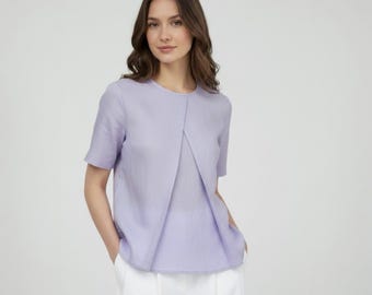 May include: A light purple short-sleeve top with a unique front design. The top is paired with white pants. The top has a round neckline and a tailored fit. The fabric appears to be a lightweight linen or similar material.