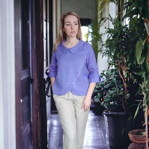 May include: A woman wearing a light purple linen blouse with a peter pan collar and white linen pants. She is standing in a hallway with plants in pots.