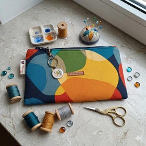 Könnte beinhalten: Eine farbenfrohe Reißverschlusstasche mit einem abstrakten Design in Blau, Grün, Orange und Gelb. Das Bild zeigt auch Garnrollen, ein Nadelkissen, eine kleine Farbpalette, dekorative Glasperlen und eine goldene Schere. Die Tasche hat einen Schlüsselanhänger mit einem runden Anhänger.