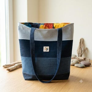 Könnte beinhalten: Eine Denim-Tragetasche mit Patchwork-Design. Die Tasche hat eine Kombination aus hell- und dunkelblauen Denim-Paneelen, mit einem grauen oberen Bereich und einem farbenfrohen gemusterten Innenraum. Sie hat zwei Denim-Träger und einen kleinen quadratischen Aufnäher mit einem Logo.