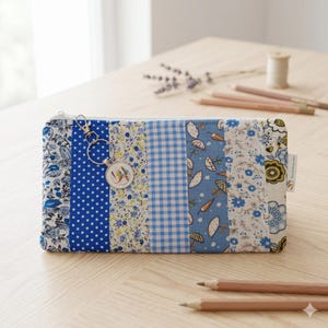 Könnte beinhalten: Ein rechteckiges Reißverschlussetui mit einem Patchwork-Design aus blauen und weißen Stoffen mit Blumen-, Punkt- und Vichy-Karomustern. Am Reißverschluss ist ein silberfarbener Schlüsselanhänger mit einem runden Anhänger und Vogelmotiv befestigt.