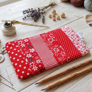 Könnte beinhalten: Ein rechteckiges Stoffetui mit einem Patchwork-Design aus roten und weißen Mustern, darunter Punkte, Streifen und florale Motive. Das Etui verfügt über einen Reißverschluss an der Oberkante und ein kleines weißes Stoffetikett an der Seitennaht.