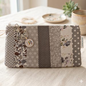 Könnte beinhalten: Eine rechteckige Reißverschlusstasche mit einem Patchwork-Design aus vertikalen Stoffstreifen in Grau-, Weiß- und Brauntönen. Die Muster umfassen Punkte, Blumenprints und geometrische Formen. Ein silberner Schlüsselanhänger mit Anhänger ist befestigt.