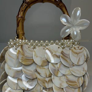 Könnte beinhalten: Eine Handtasche mit einem geschwungenen Bambusgriff und einem großen weißen Blumenakzent. Der Körper ist mit überlappenden kreisförmigen Scheiben in einem marmorierten Weiß- und Beigemuster bedeckt, akzentuiert durch eine Reihe weißer Perlen.