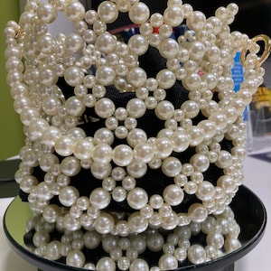 May include: A crown constructed from off-white faux pearls of different sizes. The crown sits on a black base and is reflected in a small, round mirror. The crown features a gold-coloured clasp.