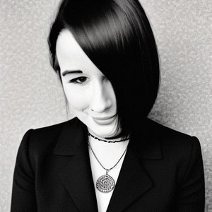 May include: Black and white portrait of a person wearing a black blazer and a necklace with a circular pendant. The person has dark hair and is smiling slightly. The pendant has an intricate design.