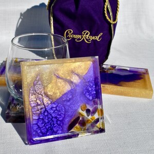 May include: Square and rectangular resin coasters with gold, purple, and clear accents. The coasters have a geode-like appearance with embedded stones. A clear wine glass and a purple bag with gold text are in the background.
