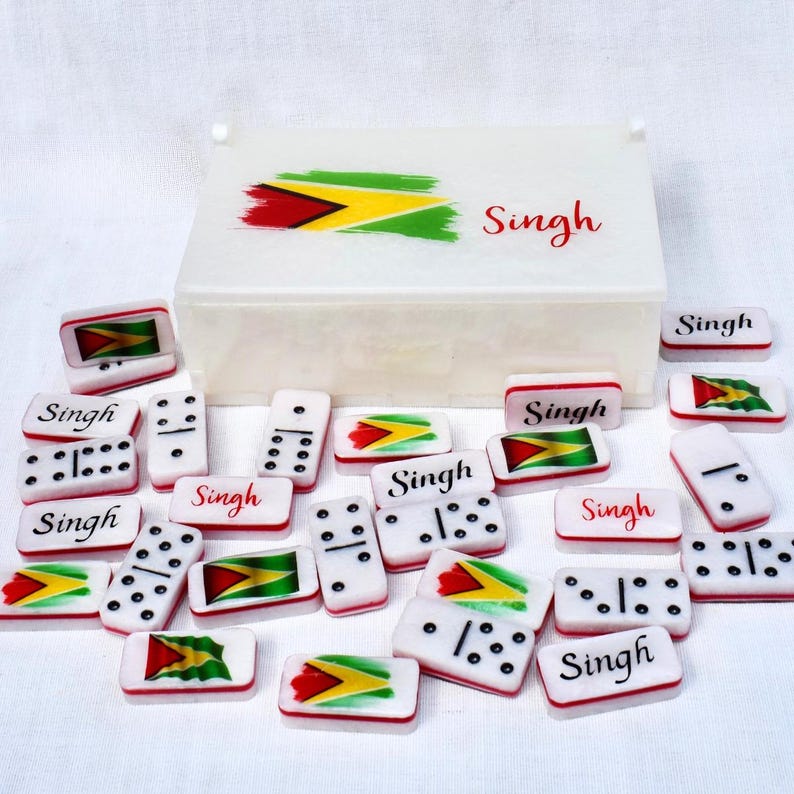 May include: A white domino set with black dots and the name "Singh" in red. Some dominoes feature the Guyana flag. The set includes a white box with the Guyana flag and the name "Singh".
