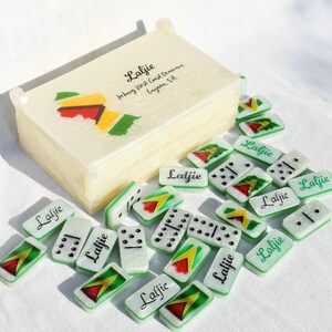 May include: A set of dominoes with a matching box. The dominoes and box are off-white with green, red, yellow, and black accents. The box has the word "Laljie" and a map of Guyana. The dominoes also have the word "Laljie".