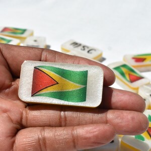 May include: A rectangular domino featuring the flag of Guyana, with red, yellow, green, and black colors. The domino is held in a hand, with other dominoes visible in the background. The dominoes appear to be made of a translucent material.