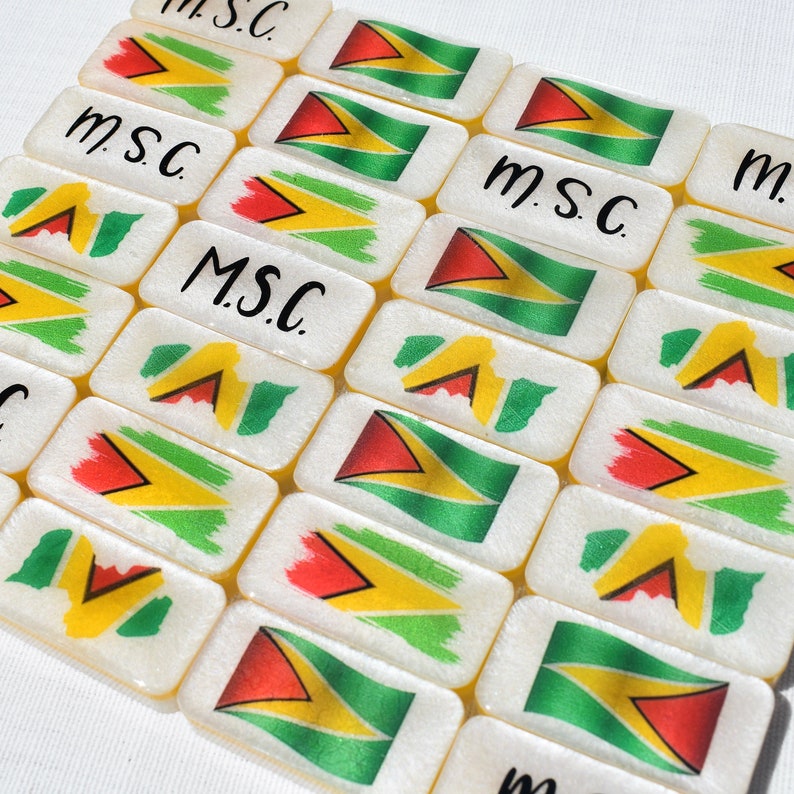 May include: Rectangular white dominoes with the flag of Guyana, featuring red, yellow, green, and black colors. The text "m.s.c." is written in black cursive on some of the dominoes. The dominoes are arranged in a grid.