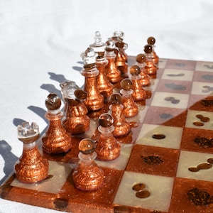 May include: A copper and white chess set with clear glass pieces. The chessboard is made of resin and has a copper glitter finish. The pieces are all in their starting positions.