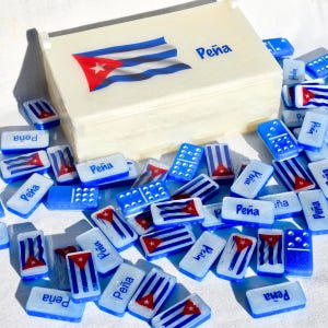 May include: A dominoes set with a cream-coloured box displaying the Cuban flag and the name "Peña". The dominoes are white and blue, with the Cuban flag and the name "Peña" printed on them. Some dominoes have dots.