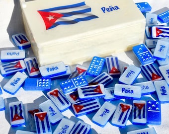 May include: A set of dominoes with a cream-colored box featuring the Cuban flag and the name "Peña". The dominoes are white and blue, with the Cuban flag and the name "Peña" printed on them. Some dominoes have dots.