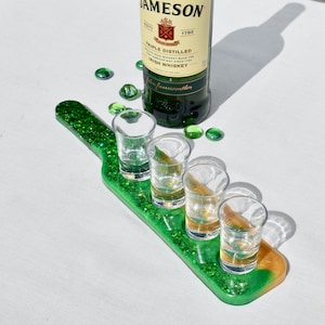 May include: A green glitter shot glass holder shaped like a bottle with four shot glasses filled with a clear liquid. The holder has a gold accent at the bottom. A bottle of Jameson Irish Whiskey is in the background.