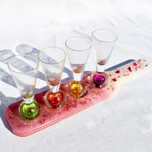 May include: A set of four clear glass shot glasses with colorful glass bases, arranged on a pink and white resin serving tray with glitter accents.