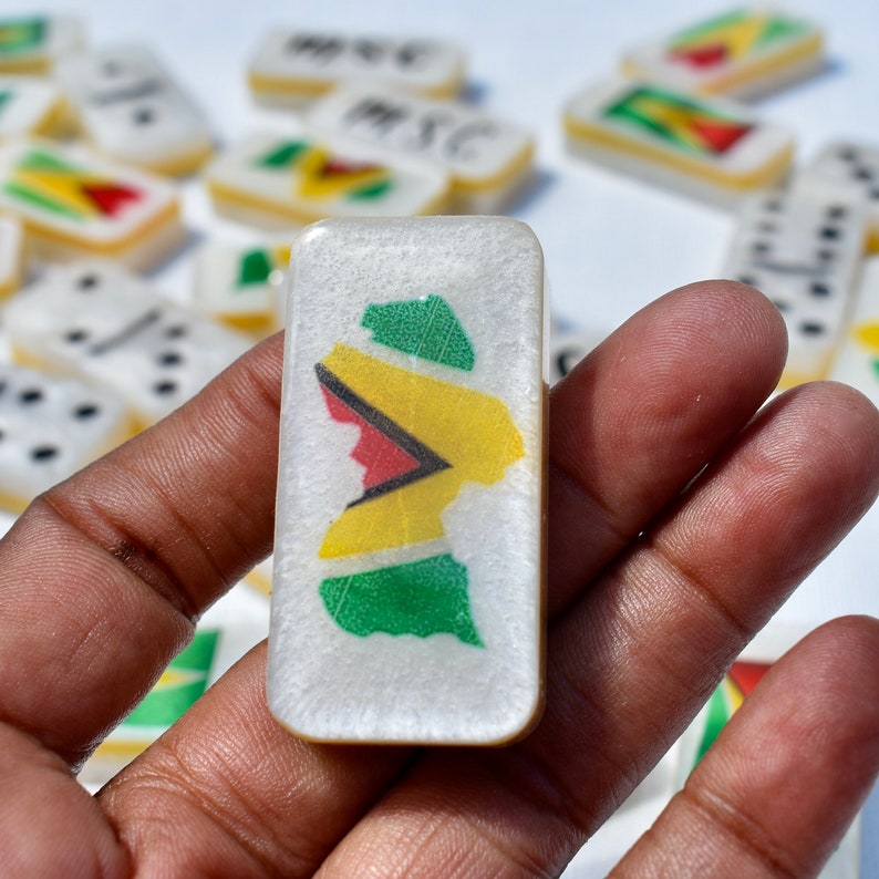 May include: A close-up of a domino tile featuring a map design with green, yellow, and red colors. The domino is held in a hand, with other dominoes visible in the background. The dominoes appear to be part of a game set.
