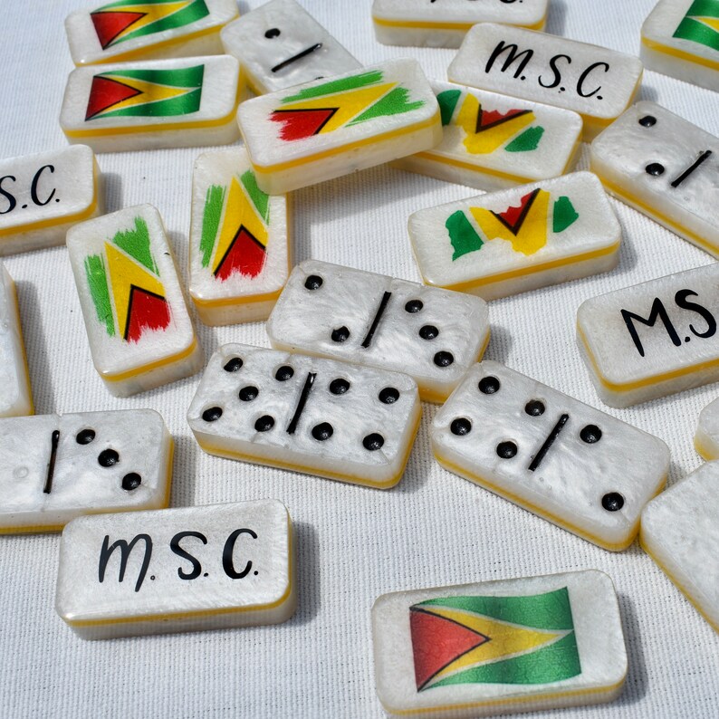 May include: A collection of white dominoes with black dots and the flag of Guyana. Some dominoes have the letters "m.S.C." written on them. The dominoes have a yellow border.
