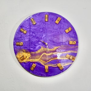 May include: Round wall clock with a purple and gold resin design. The clock face features gold-colored dominoes as hour markers and gold clock hands. The clock is mounted on a white wall.