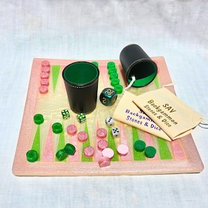 May include: A backgammon set featuring a light pink board with green and pink triangular points, two black leather dice cups with green interiors, green and pink game pieces, dice, and a beige drawstring bag labeled SAV Backgammon Stones & Dice.