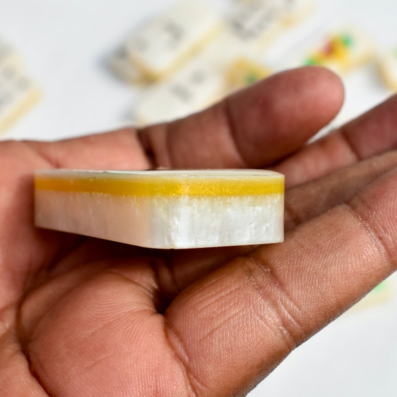 May include: A rectangular, layered object with a white base, a gold-colored middle section, and a clear top layer. The object is held in a hand, with other similar objects visible in the background. The object appears to be a component of a game.