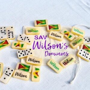 May include: A collection of ivory-colored dominoes with black dots and the word "Wilson" printed on them. Some dominoes feature a flag design with red, yellow, and green colors. A drawstring bag with the text "SAV Wilson's Dominoes" is also present.